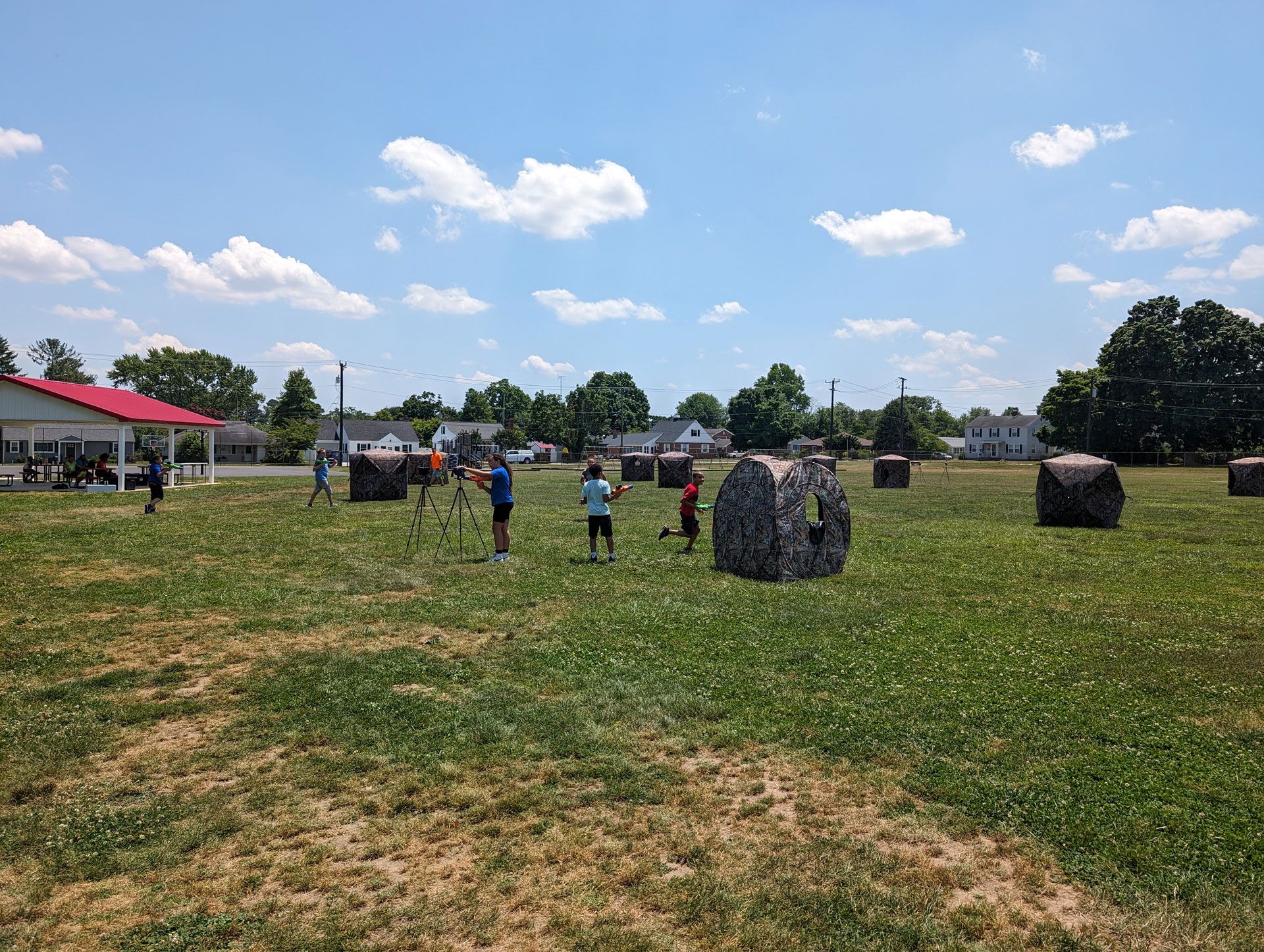 Four individuals playing paintball in a grassy field with colorful inflatable bunkers, holding markers and wearing shorts.