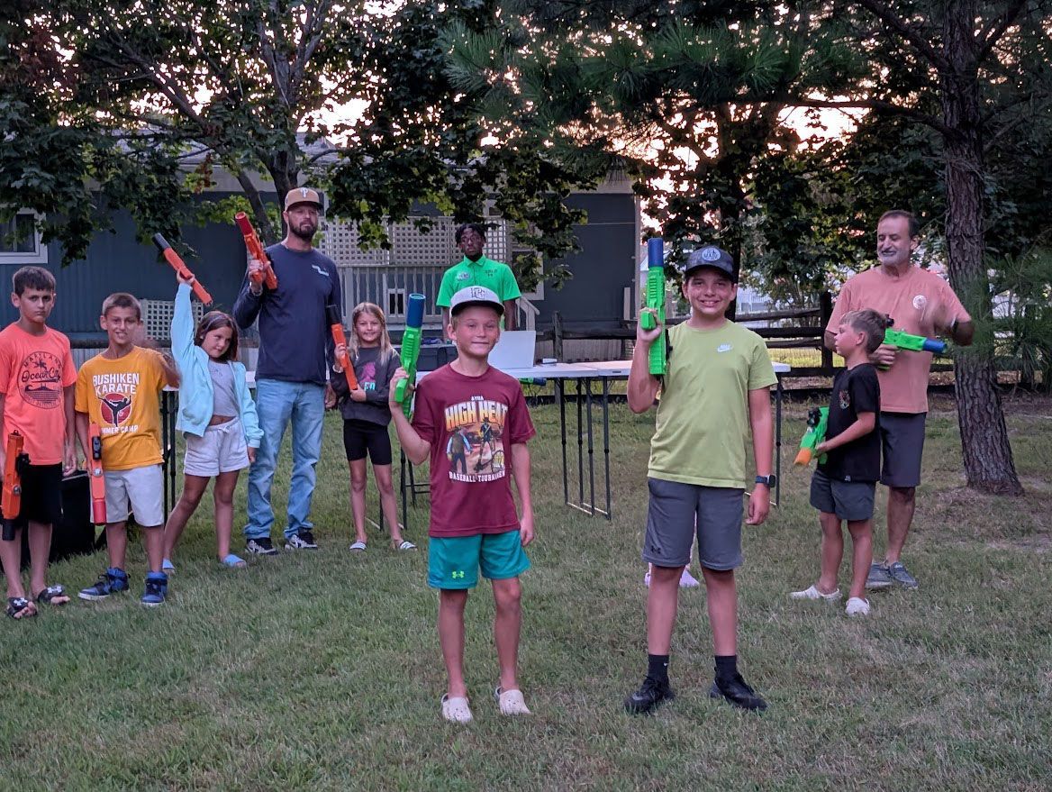 A group of children and adults standing on a grassy lawn outdoors, smiling while holding plastic water blasters.