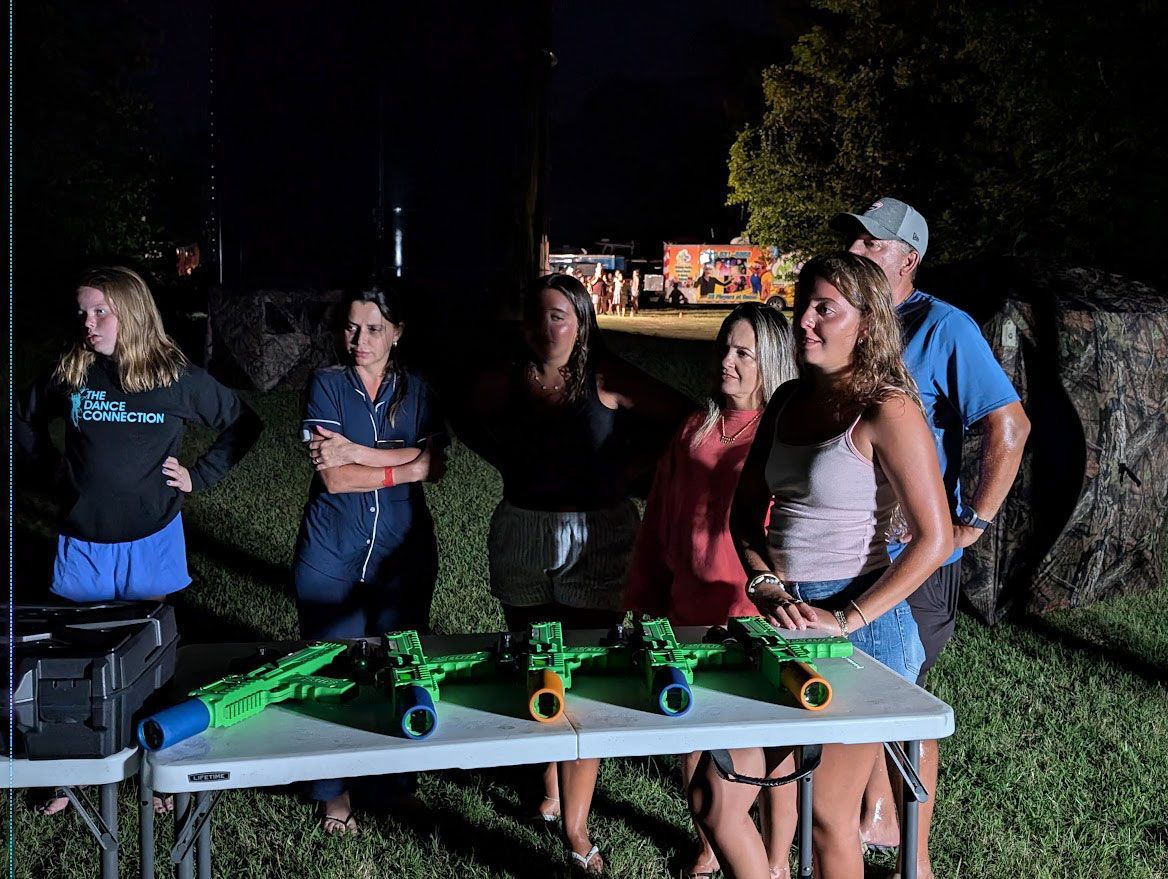 A group of people standing behind a table at night with several green laser tag-style blasters lined up.