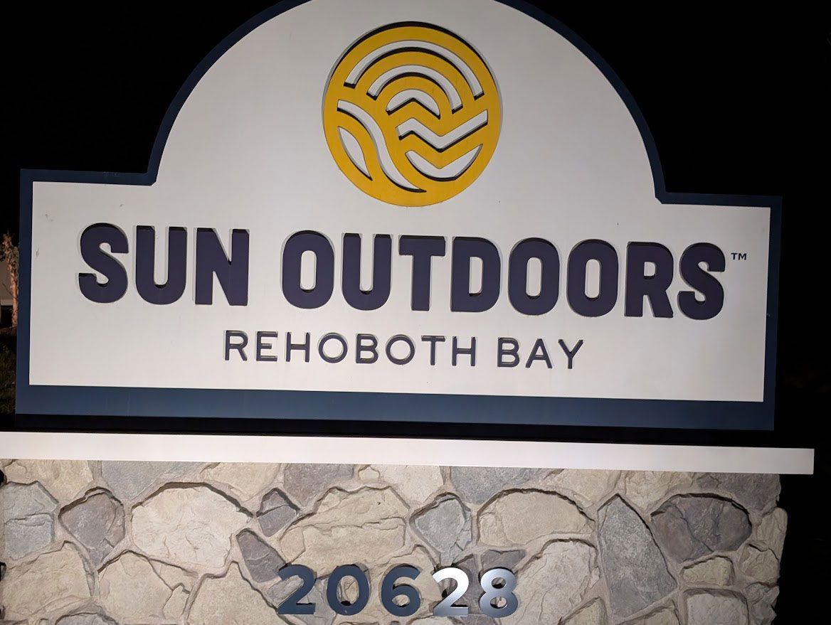 Sign for Sun Outdoors Rehoboth Bay featuring a yellow sun logo on a white arch over stone base with address 20628.