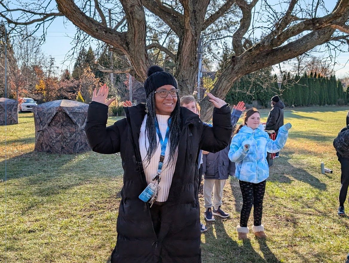 A person in a black coat and hat with hands raised stands outdoors in a sunny field with people behind them.