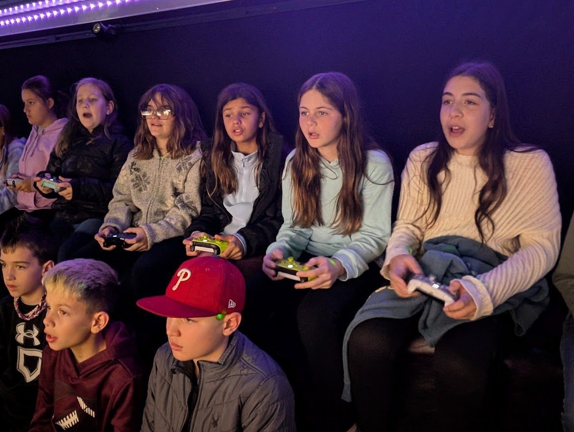 A group of people sitting indoors, focused and holding game controllers while gaming in a dimly lit room.