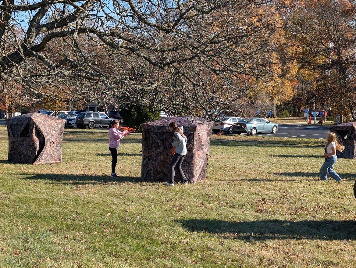 People in casual clothing play a game among several camouflage-patterned pop-up tents on a grassy field.