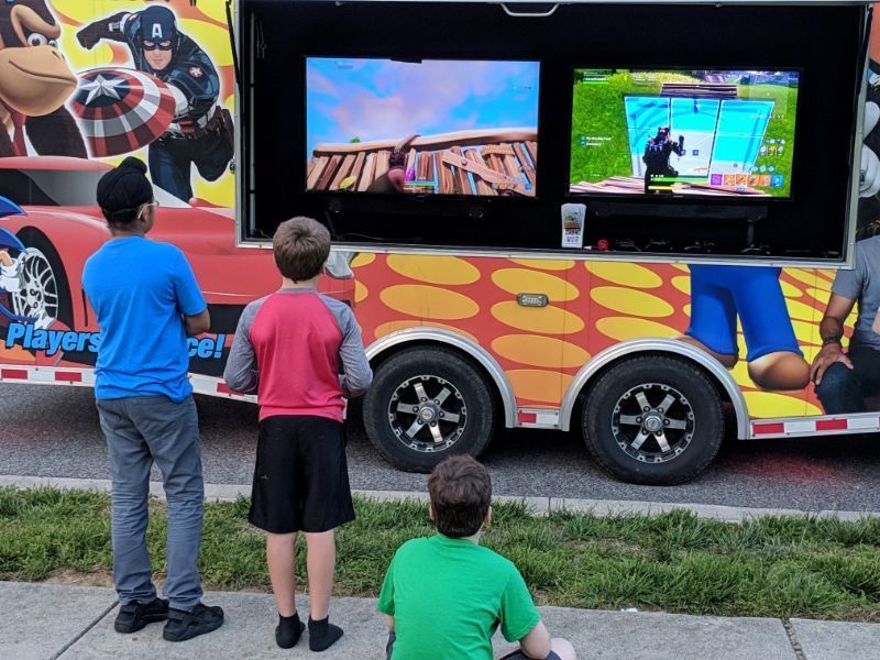 Three children watch games on large monitors mounted to the side of a colorful gaming trailer parked on a sidewalk.