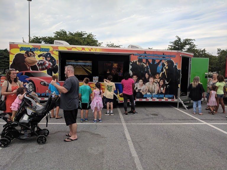 People play video games inside a mobile arcade trailer parked in an outdoor lot on a sunny day.
