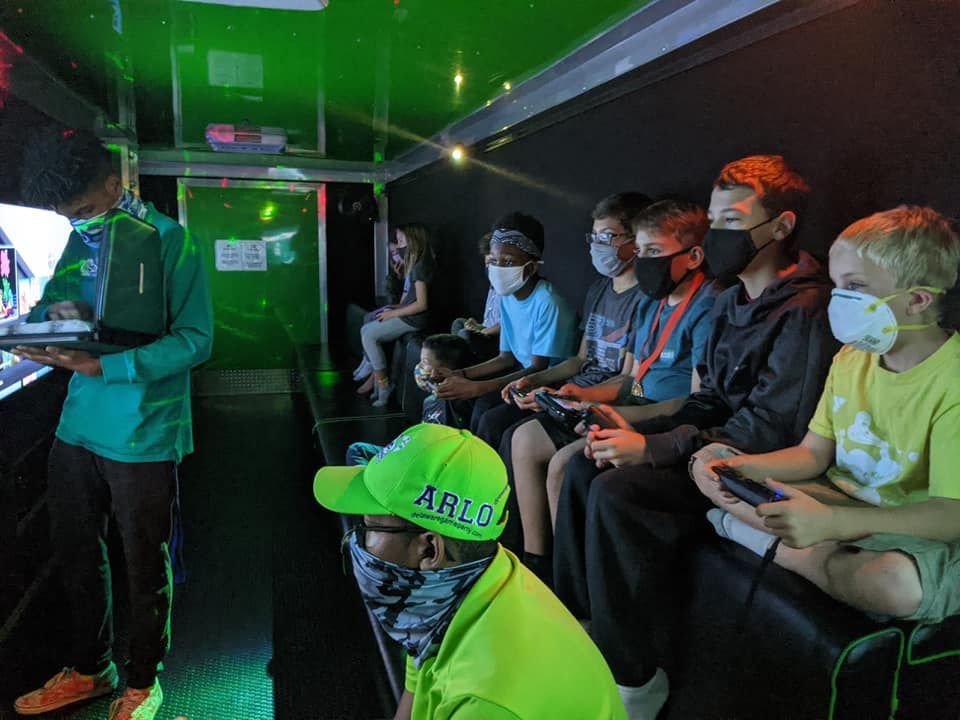 A group of masked individuals play video games inside a mobile trailer with green lighting.