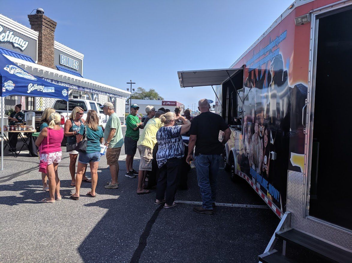 People wait in line at a food truck parked on a sunny paved lot next to a building with a blue awning.