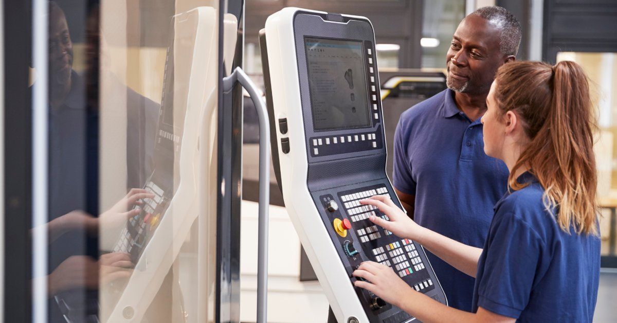 Two workers wearing blue shirts using a terminal to enter a variety of commands into a CNC machine.