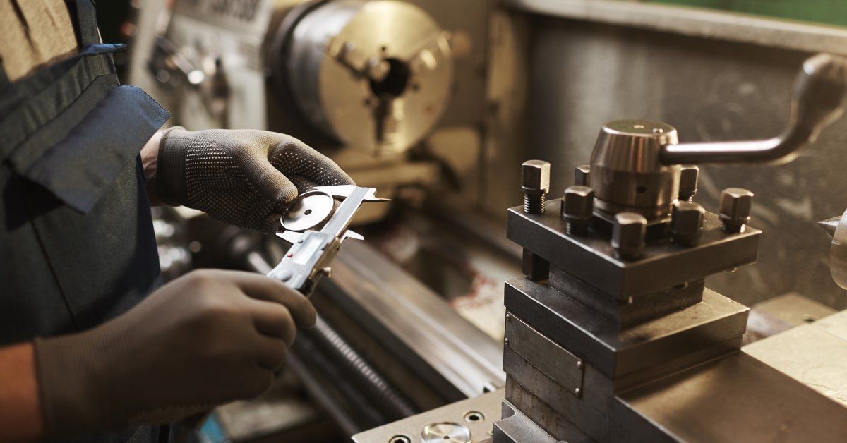 A worker in an industrial setting wearing a protective apron and gloves using a tool to measure a metal component.