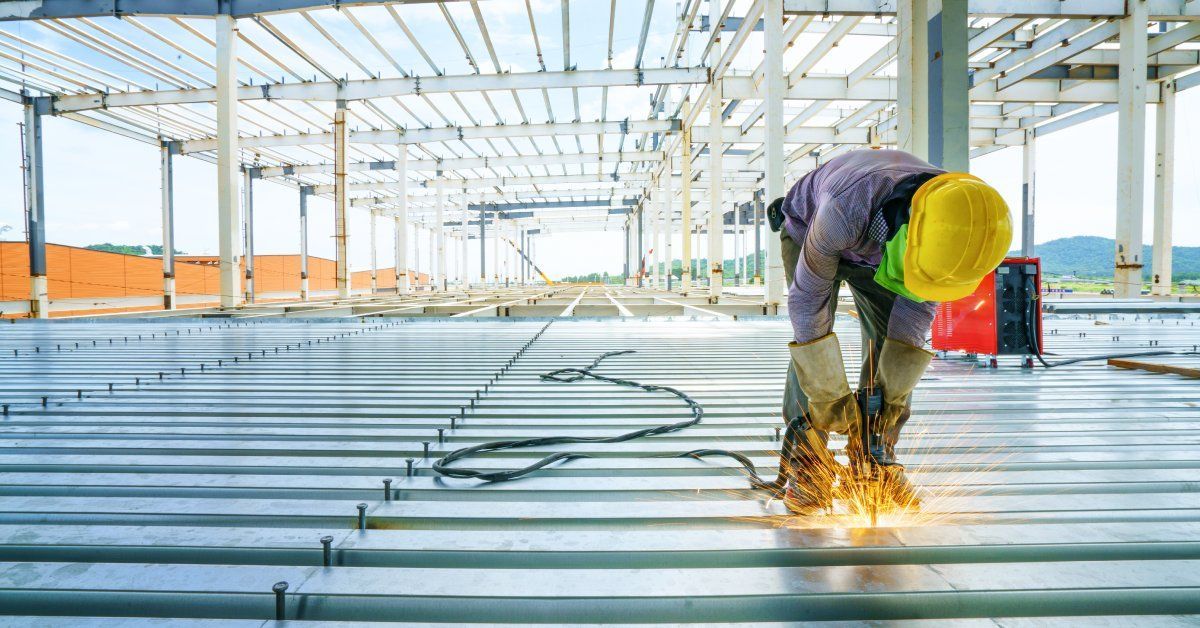 A worker wearing a yellow hard hat leaning over a metal structure using a welder to join parts.