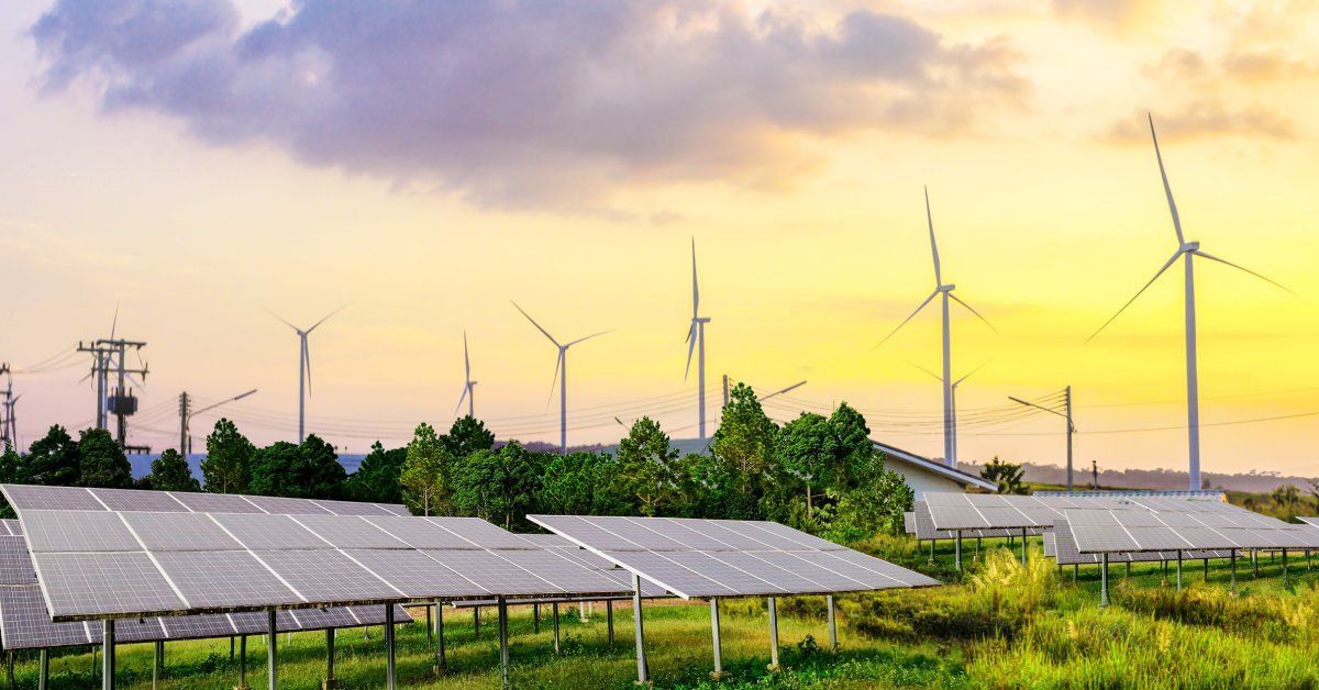 Solar panel installations sitting in the grass with wind turbines and power lines in the background.