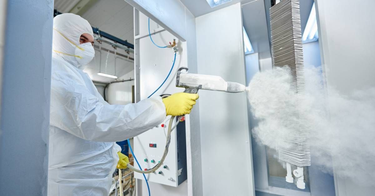 An industrial worker wearing a protective bodysuit and face mask using a spray gun to apply coating.