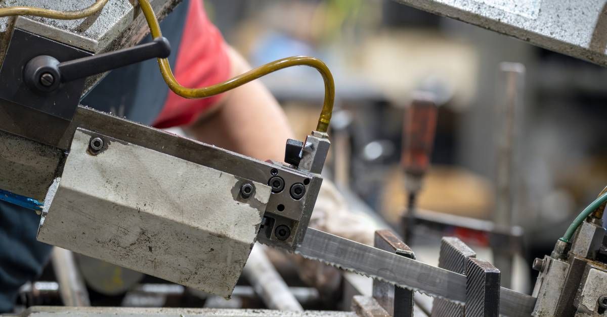 A worker stands next to an automated machine and uses a saw blade to cut through a piece of metal.