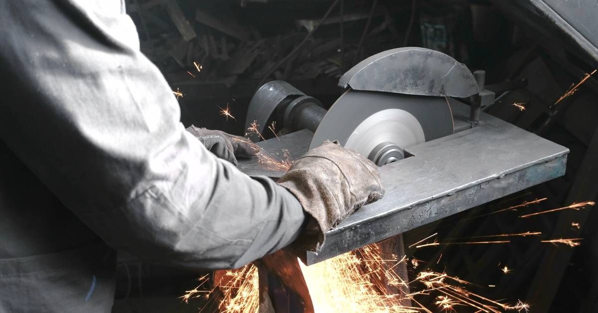 A worker wearing a long-sleeve shirt and gloves feeds a piece of metal through a circular saw blade.