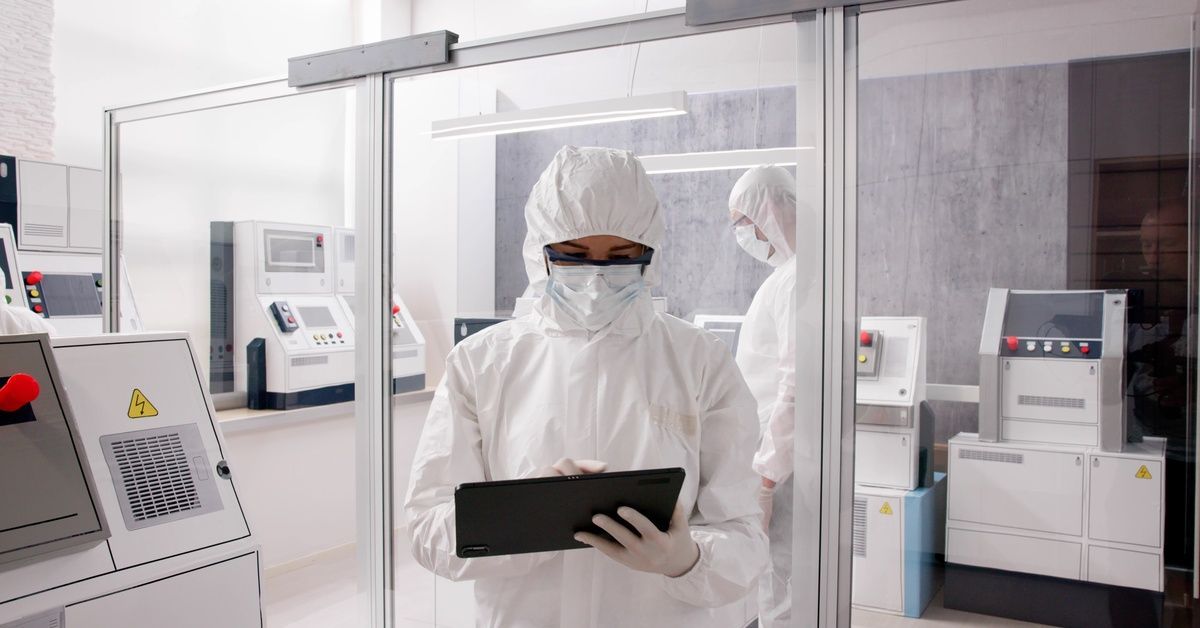 Two people wearing white coveralls, gloves, masks, and safety goggles while standing in a clean room to check various machines.