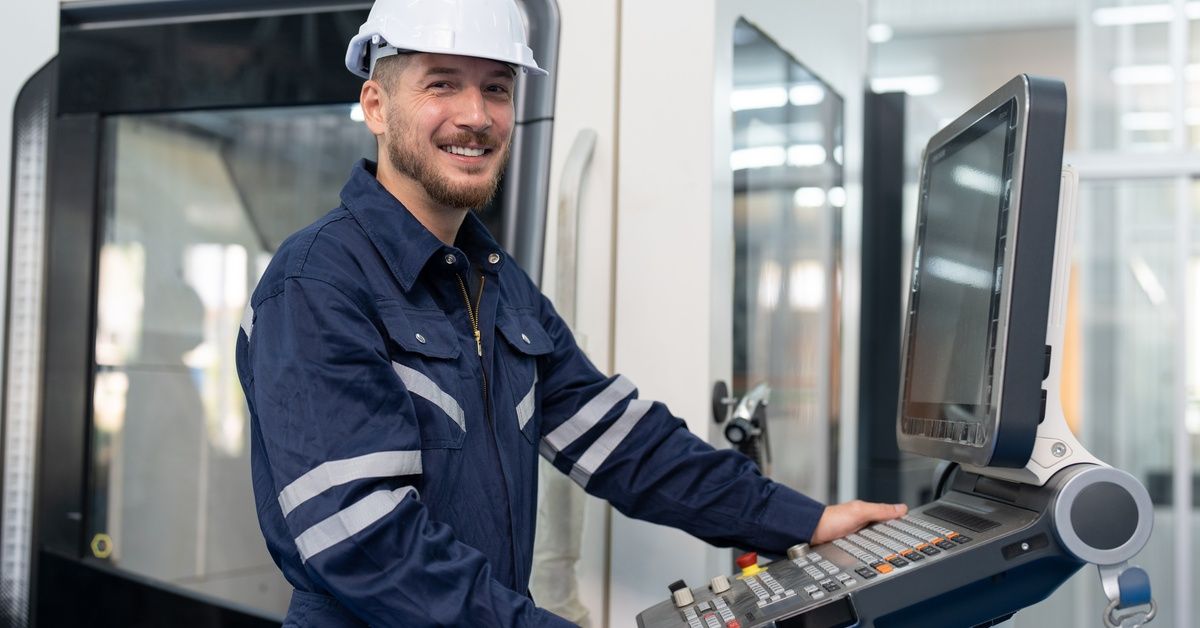 A man in a blue jumpsuit and white hard hat, standing next to a control panel for a CNC machine.