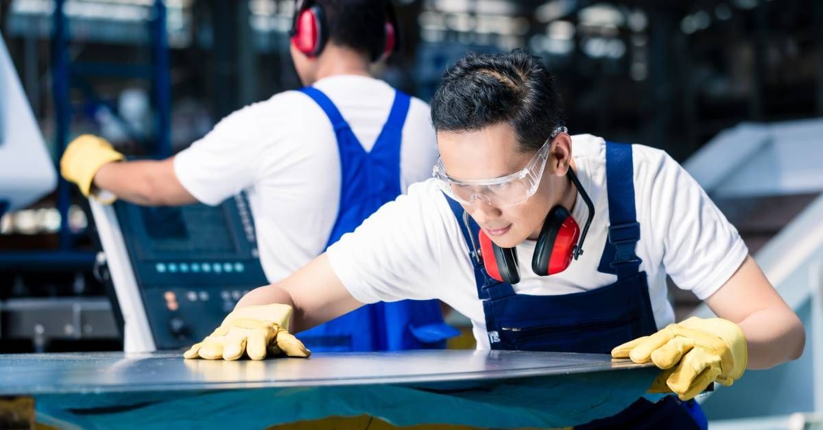 A worker in a metal manufacturing industry looking carefully at a piece of sheet metal with another worker behind him.