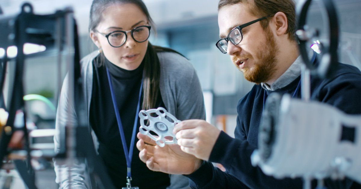 A business man and woman surrounded by equipment, discussing a prototype that they created.