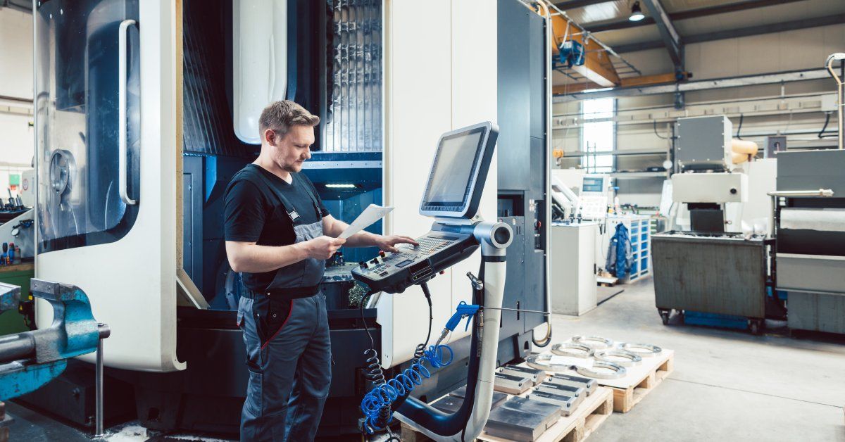 A worker on a manufacturing floor holding a piece of paper and entering commands into a terminal.