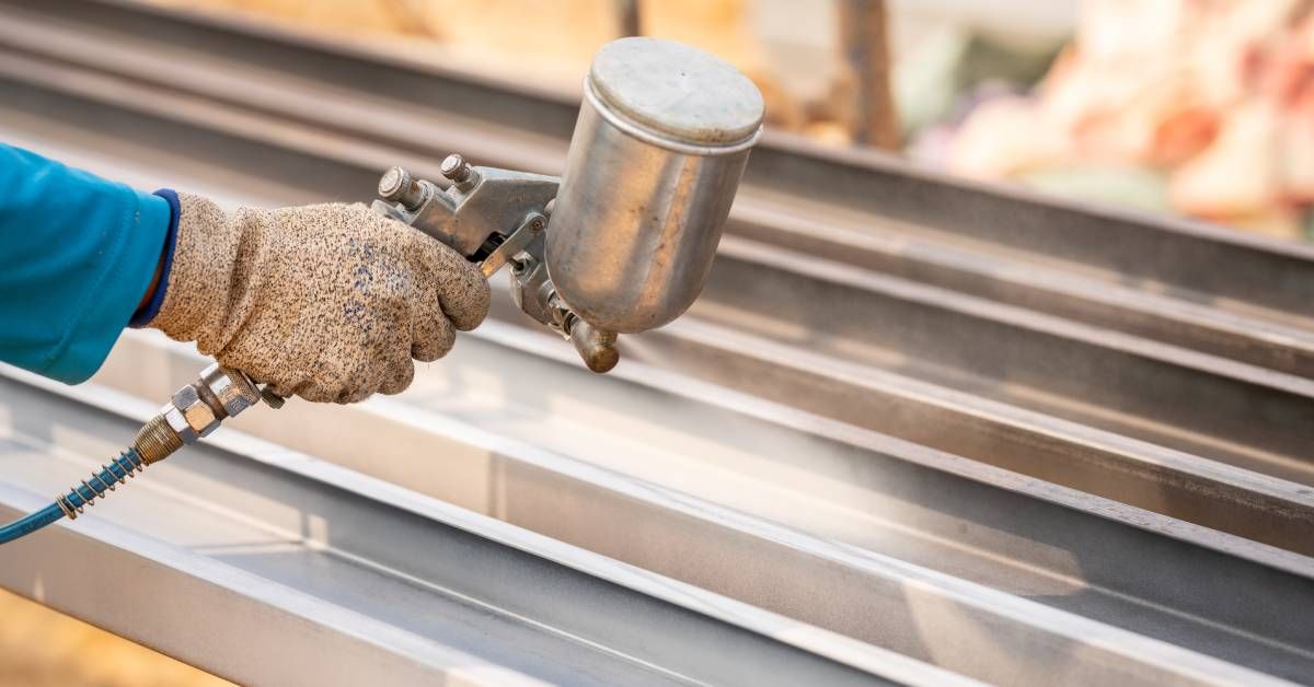 A close-up of a worker’s hand using a spray gun to apply an industrial coating onto rows of metal beams.