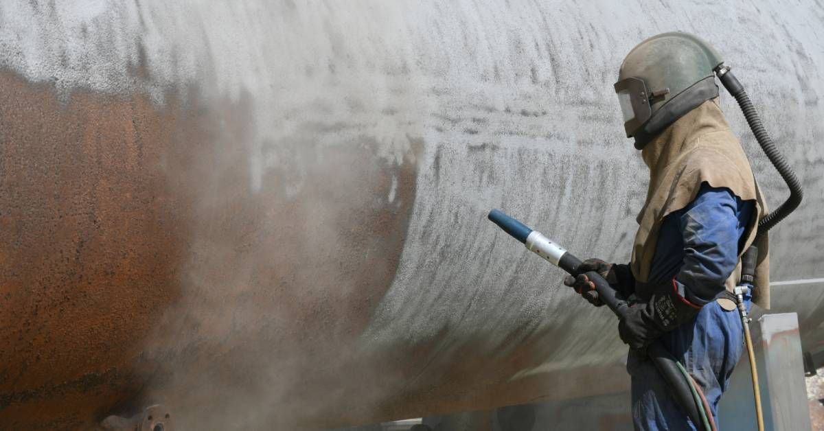 A worker in an industrial setting wearing a face mask and respirator while spraying onto a tank.