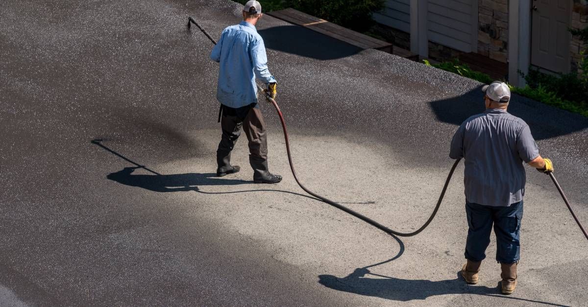 Two workers standing outside using a hose attached to a spray gun to apply a protective coating onto asphalt.