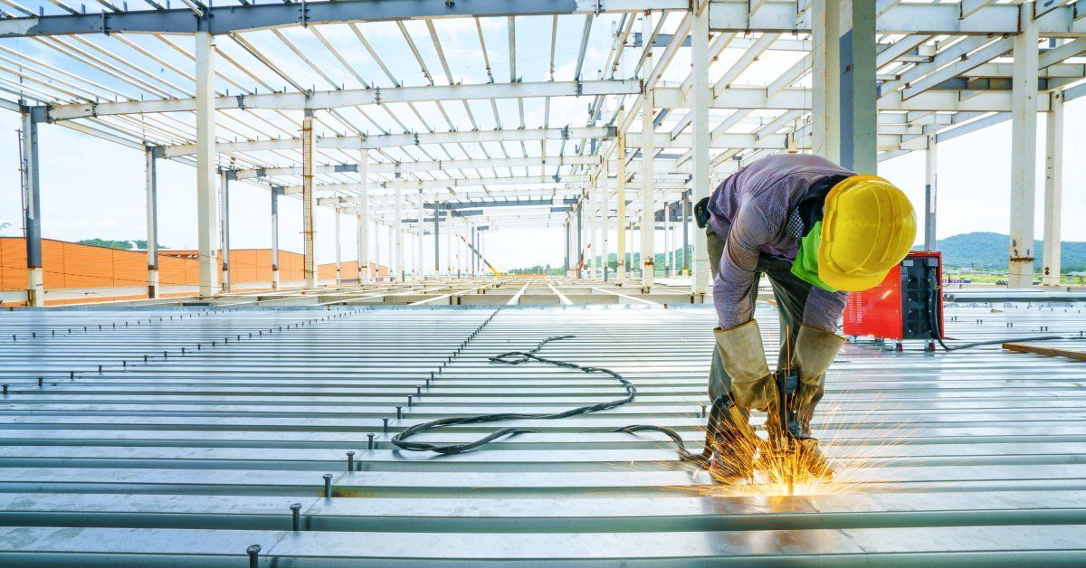 A worker wearing a yellow hard hat and protective gloves kneeling over while fabricating metal.