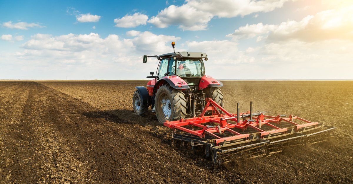 A farmer operates a red tractor on a dirt field, cultivating the land in front of a cloudy sky.