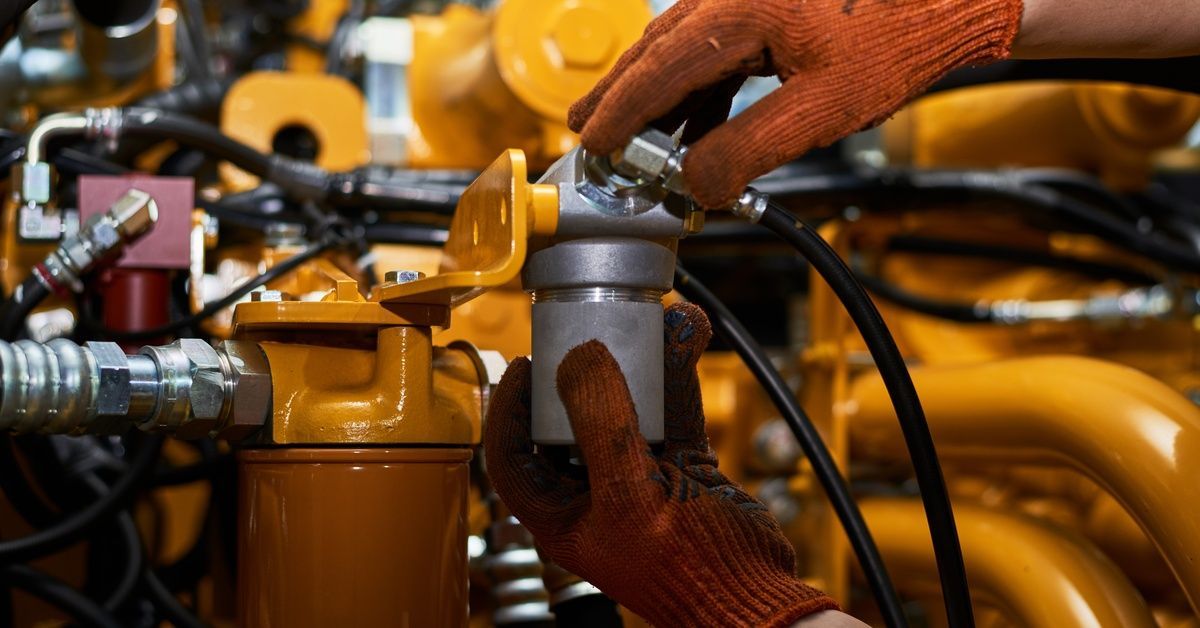 A technician wearing dark orange gloves holds onto a metal component of a yellow industrial machine.