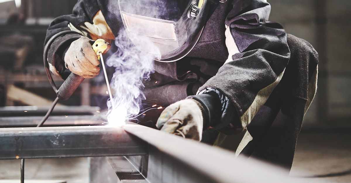 A factory worker wearing dark protective clothing and a welding helmet uses a welding rod to connect two metal pieces.
