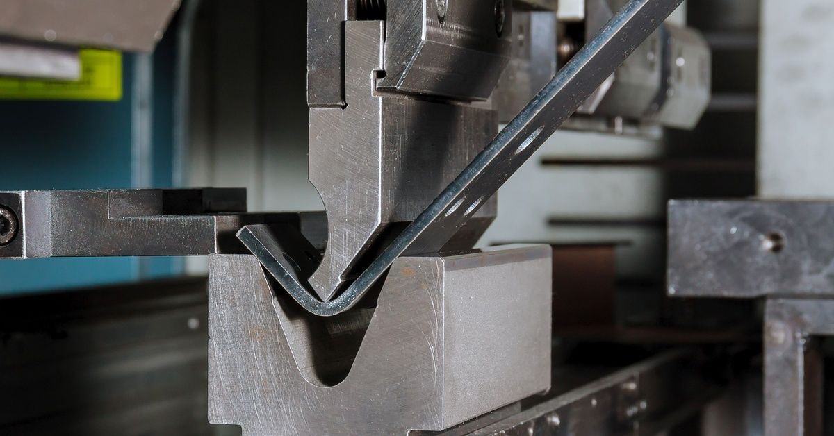 A hydraulic press machine uses force to bend a piece of metal with drilled holes to create a 90-degree angle.