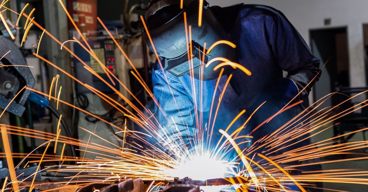 A welder wearing protective gear works in a factory welding on a work surface, creating sparks.