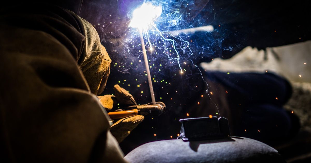 A close-up of a welder using a stick and welding gun to fuse two pieces of metal above their head, creating bright light.