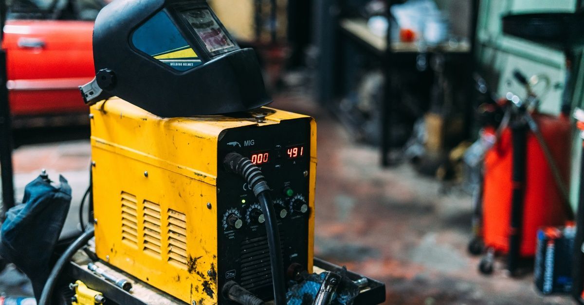 A yellow welding machine box with a welder's protective mask sitting on top in a garage with a red car behind.
