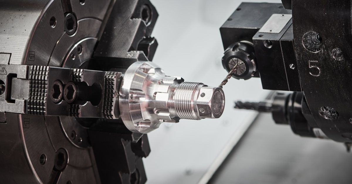 A CNC lathe machine holds a carved piece of stainless steel, removing the material with a tool.