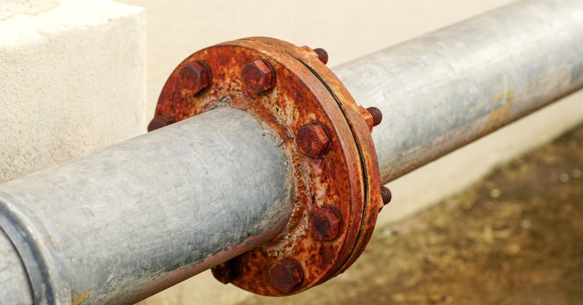 A close-up of a steel water pipeline that has significant rust where two pipes meet, secured by rusted bolts.