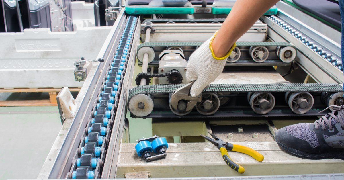 A worker wearing gloves uses a wrench to adjust a wheel under a conveyor belt in a factory.