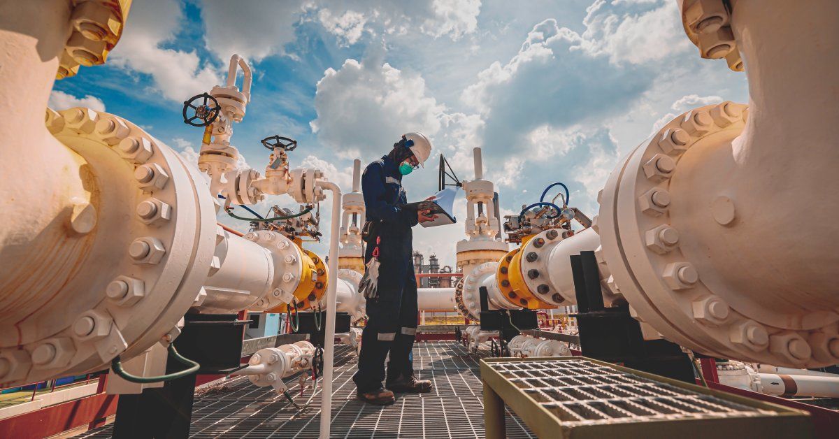 A man holds a clipboard while performing an inspection near large steel pipes at an oil refinery.