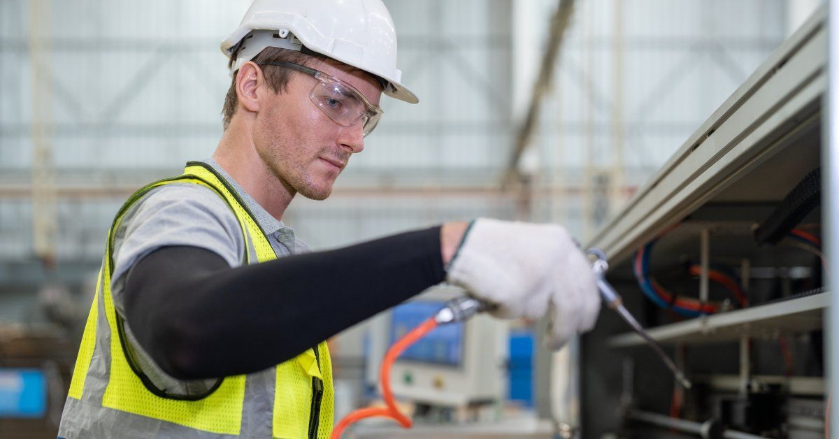 A male worker wearing a safety vest uses an air compressor to clean the inside of a machine.