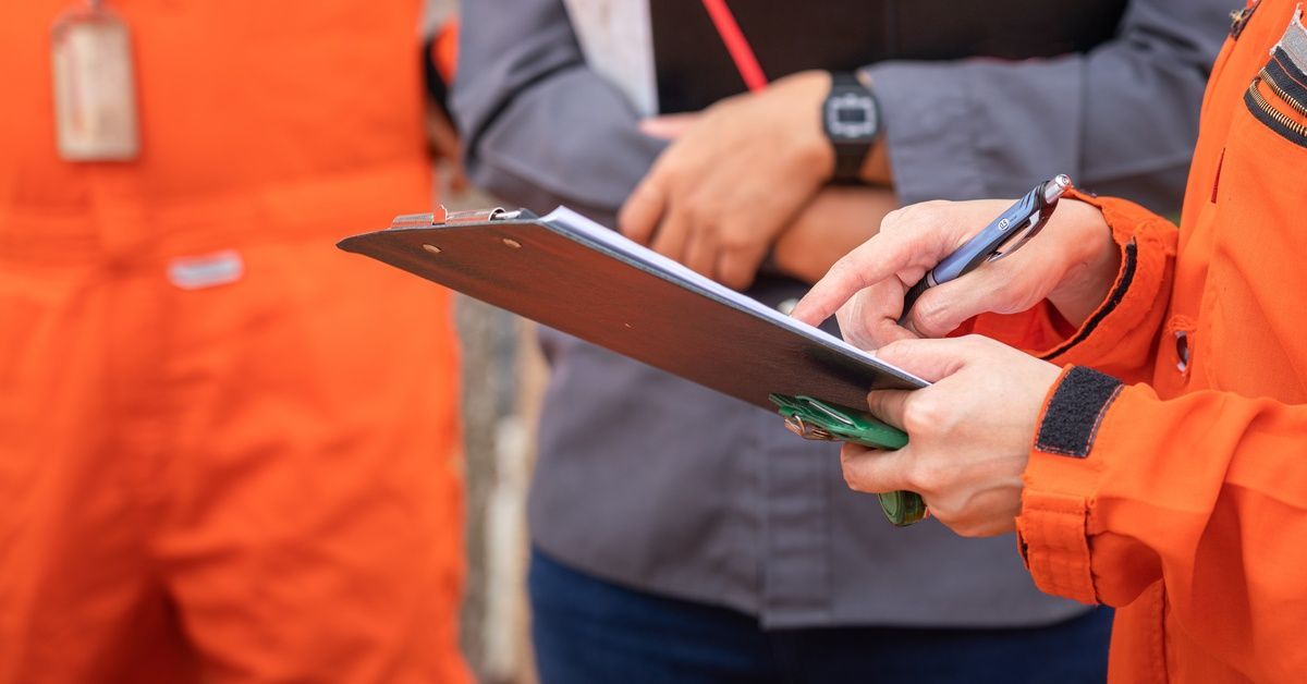 A person wearing an orange jumpsuit holds a clipboard pointing at something for two other people.