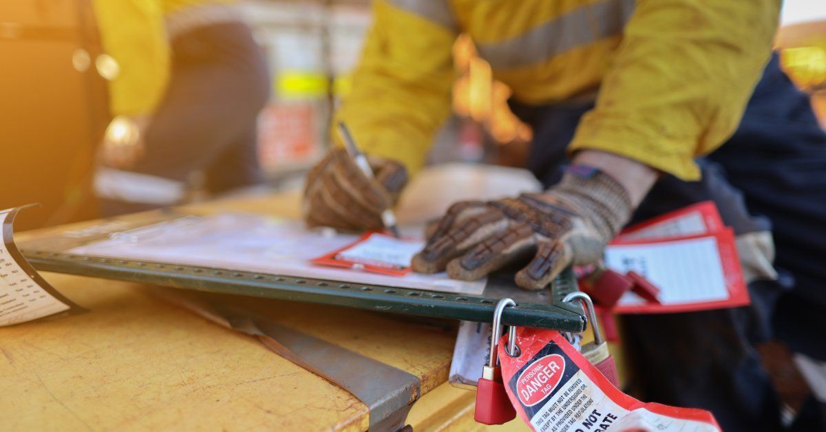 A worker wearing a yellow jacket writes on a flat surface that has locks with attached danger tags.