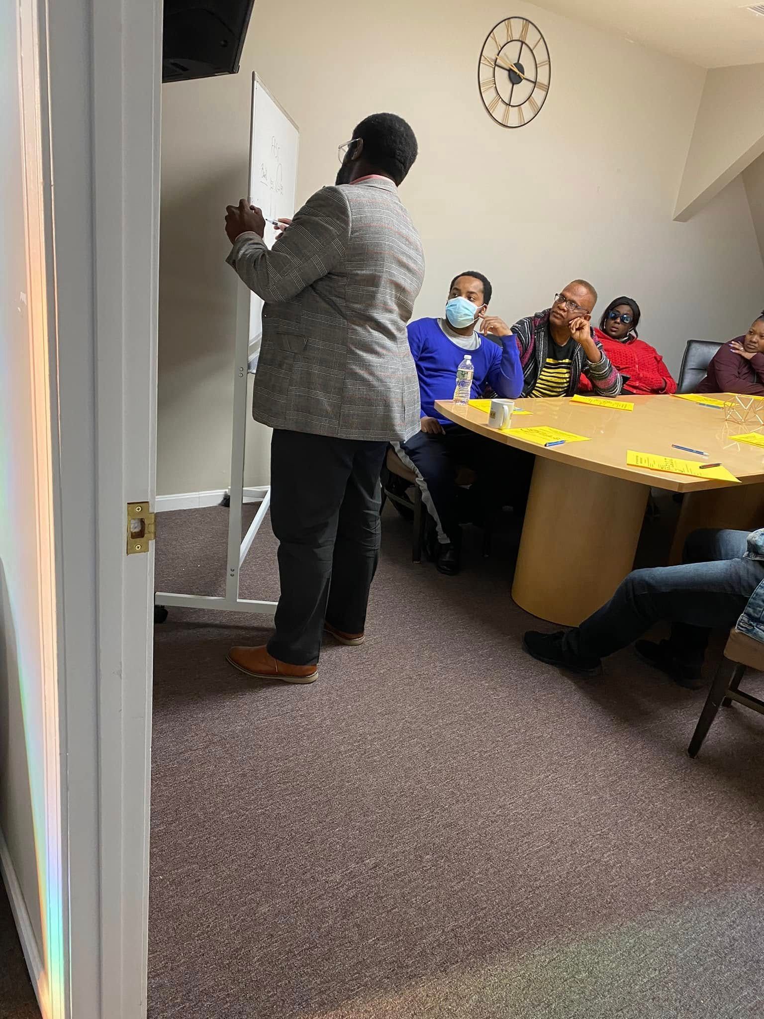 Person speaking beside a whiteboard in a small meeting room while others sit around a table.