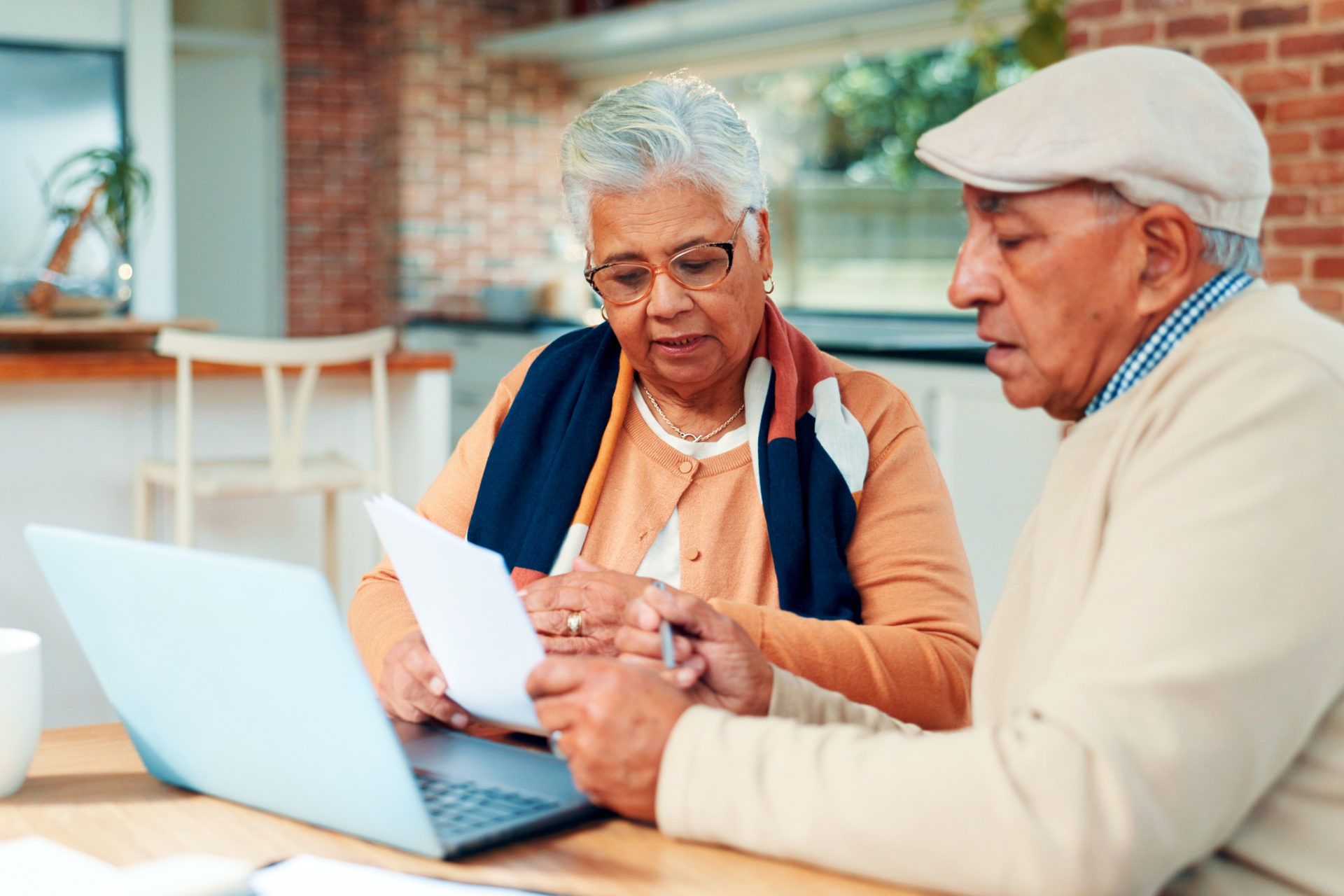 Two seniors reviewing a document at a table with a laptop in a bright home setting