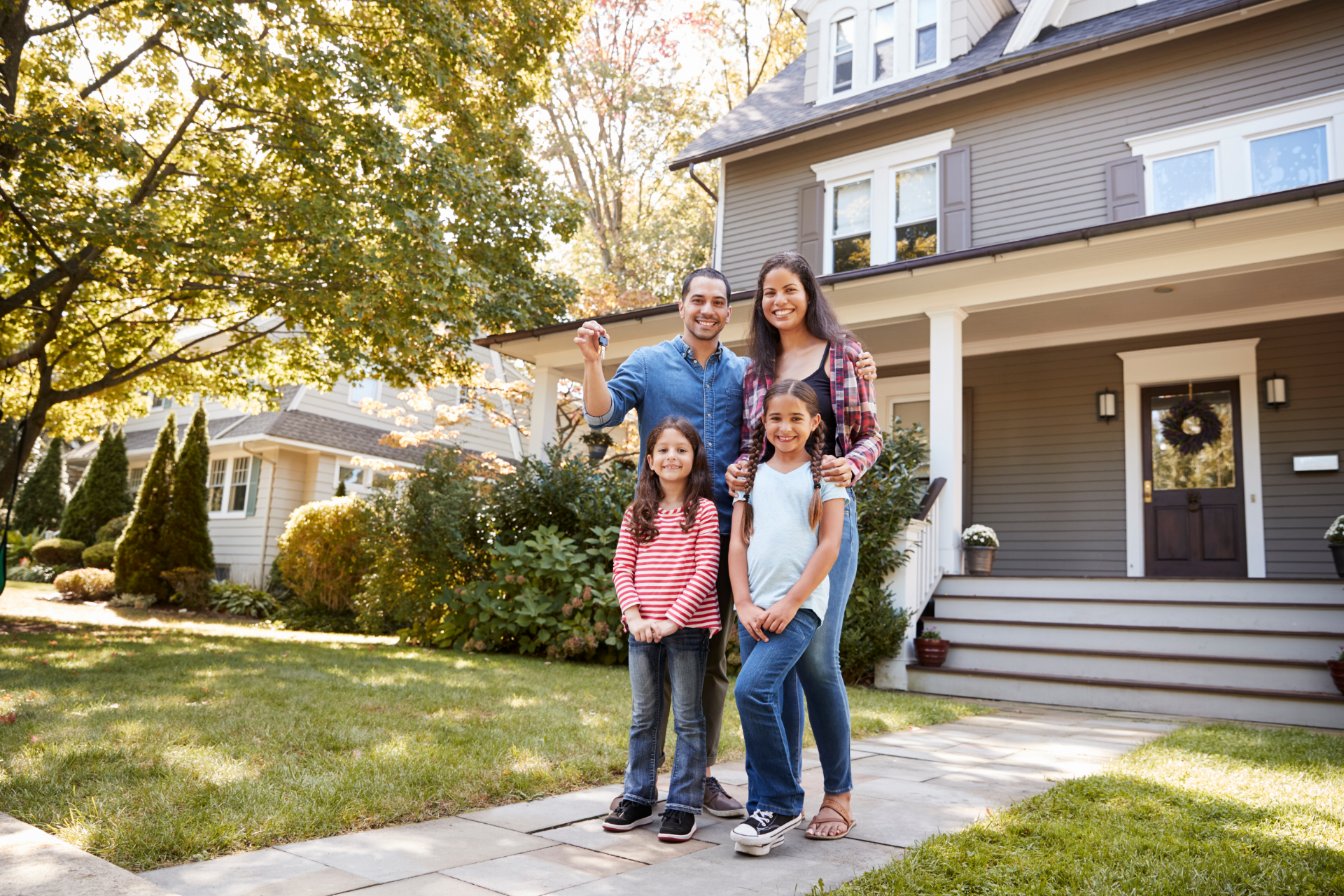 Four people standing smiling on a front walkway outside a suburban house with autumn trees.