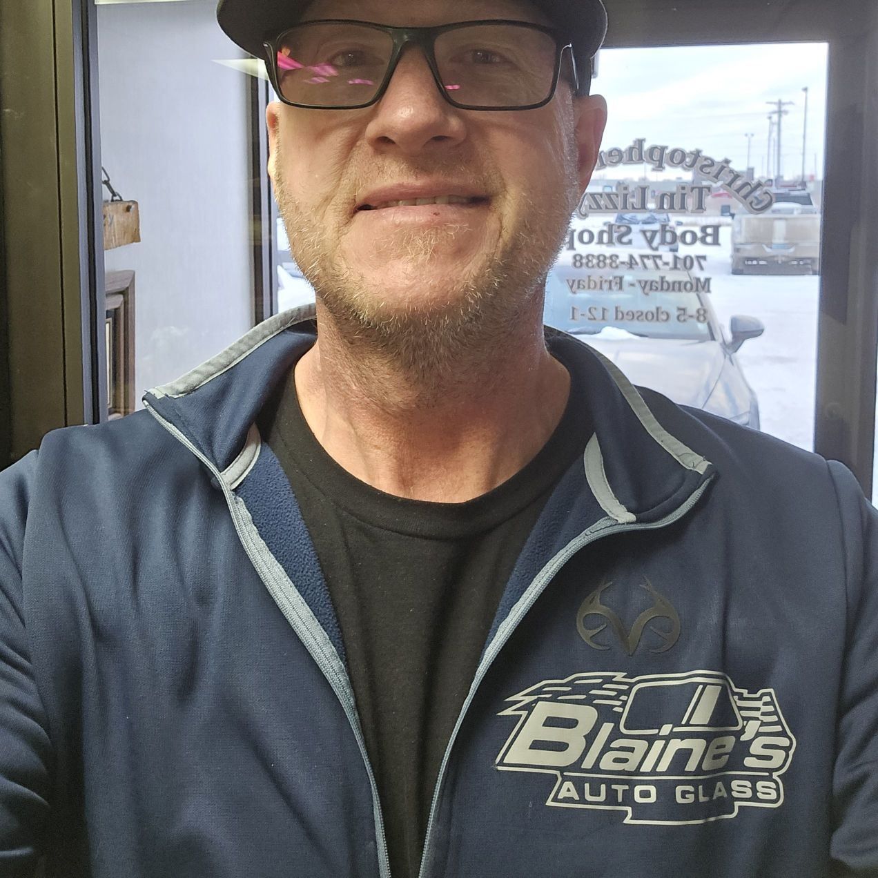 Man in glasses and jacket smiling, near a window. Jacket has 