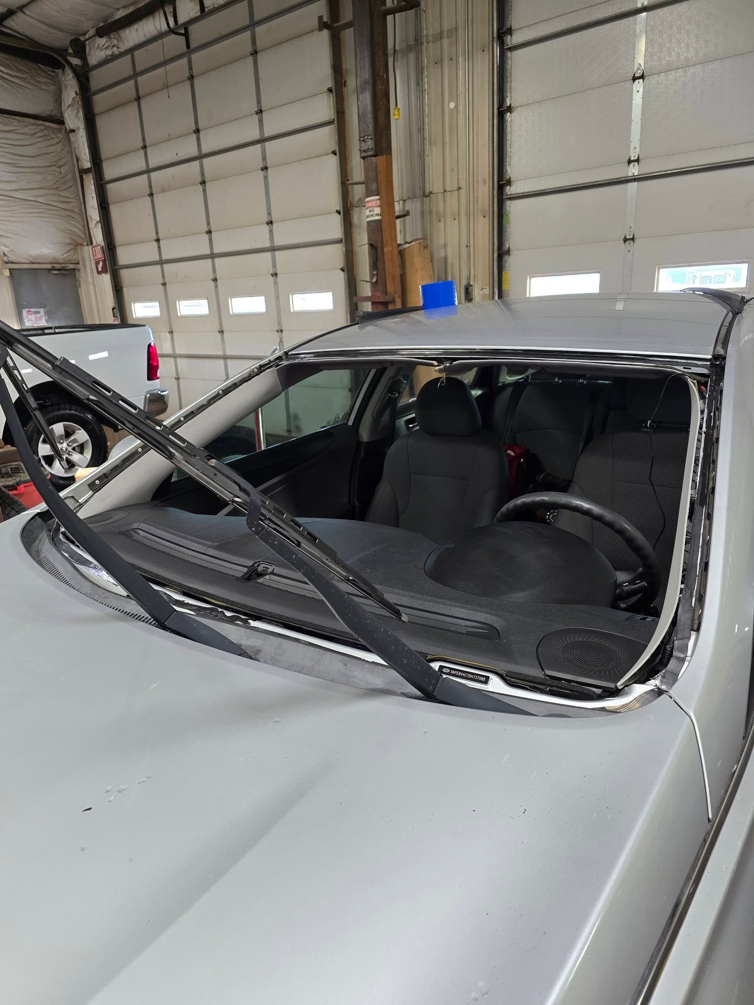 Silver car with a severely damaged windshield and windshield wipers up in a garage setting.