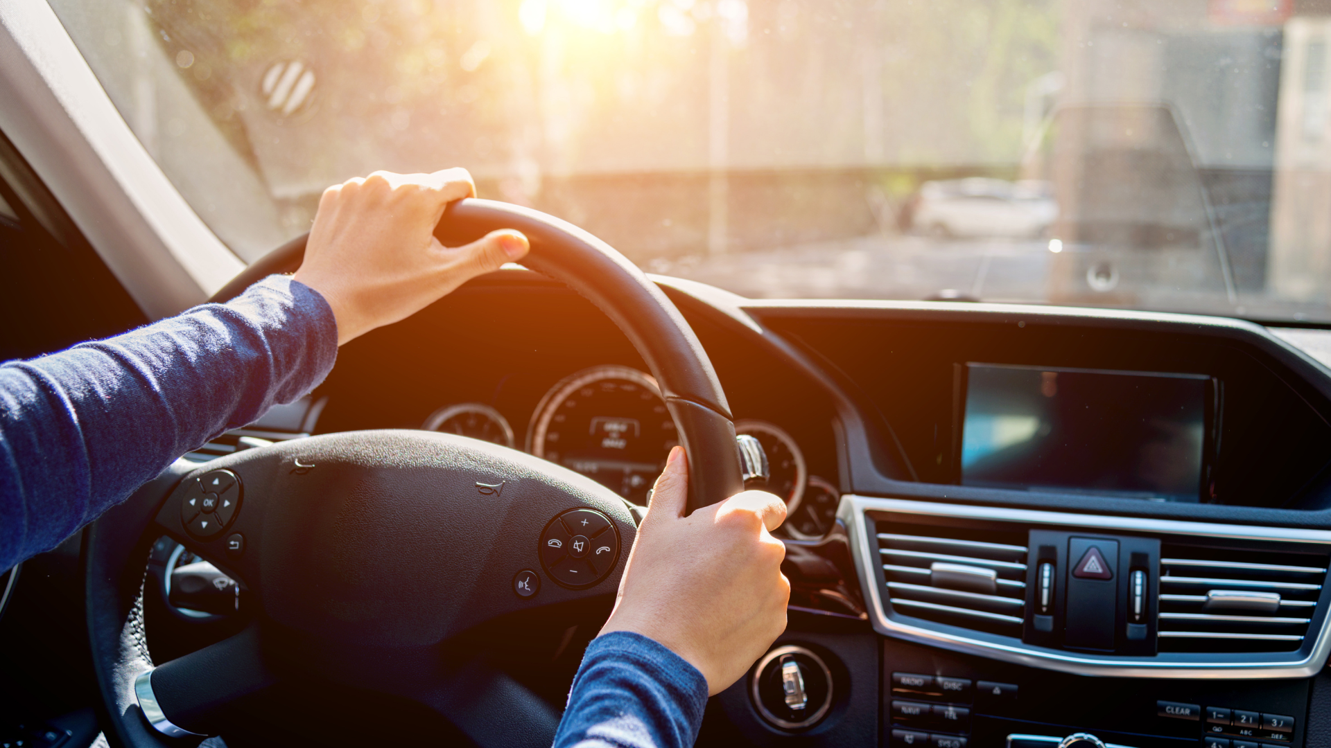 Hands gripping a car steering wheel; bright sunlight shines through the windshield.