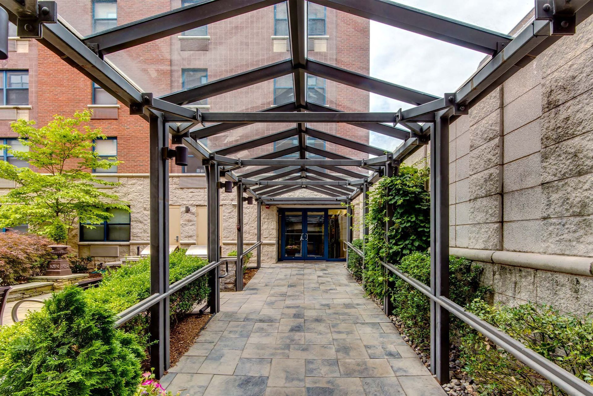 Covered walkway with glass roof, leading to blue double doors. Stone path, metal supports, and greenery on either side.