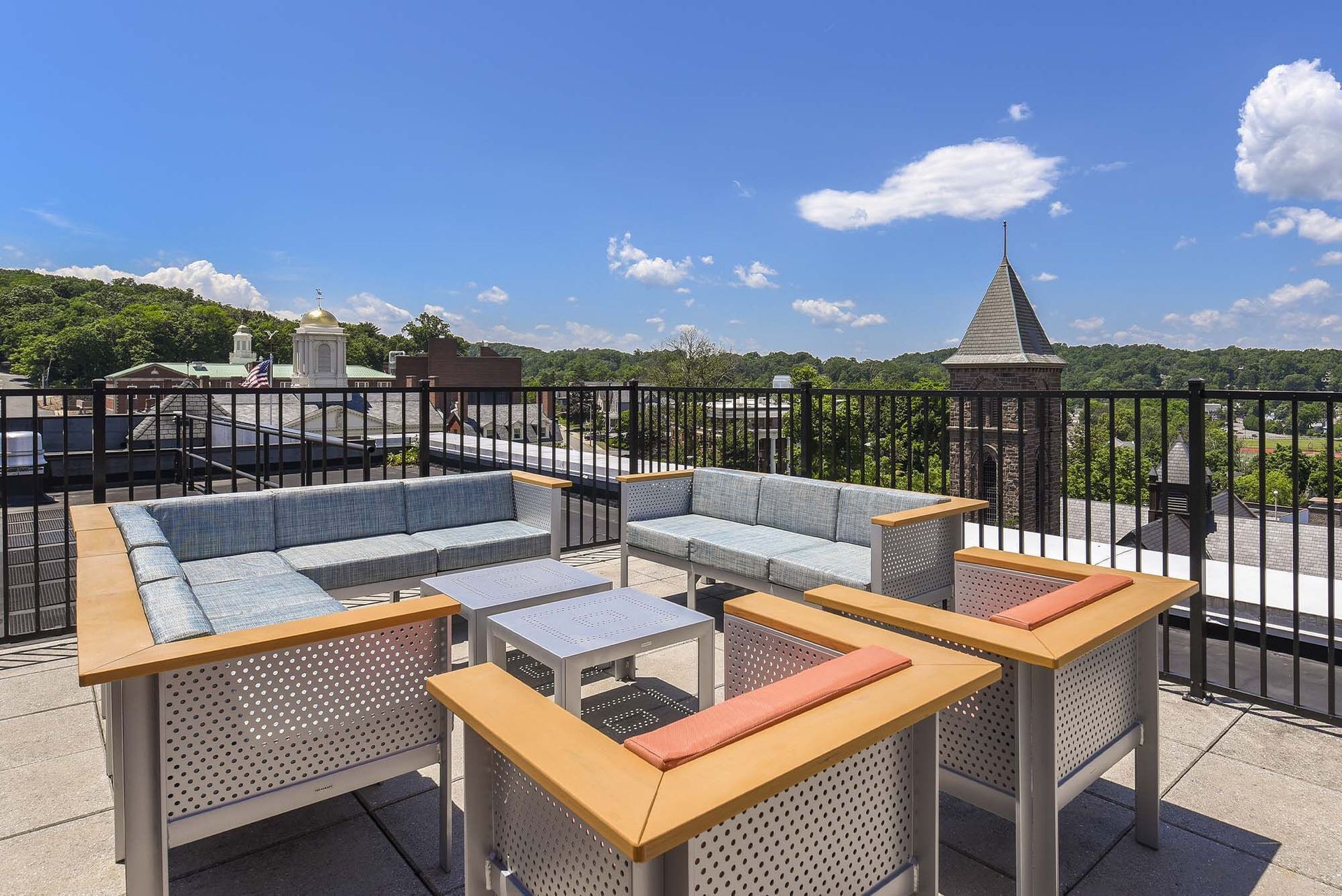 Rooftop lounge with light blue sofas, coffee tables, and a metal fence, overlooking a cityscape under a blue sky.