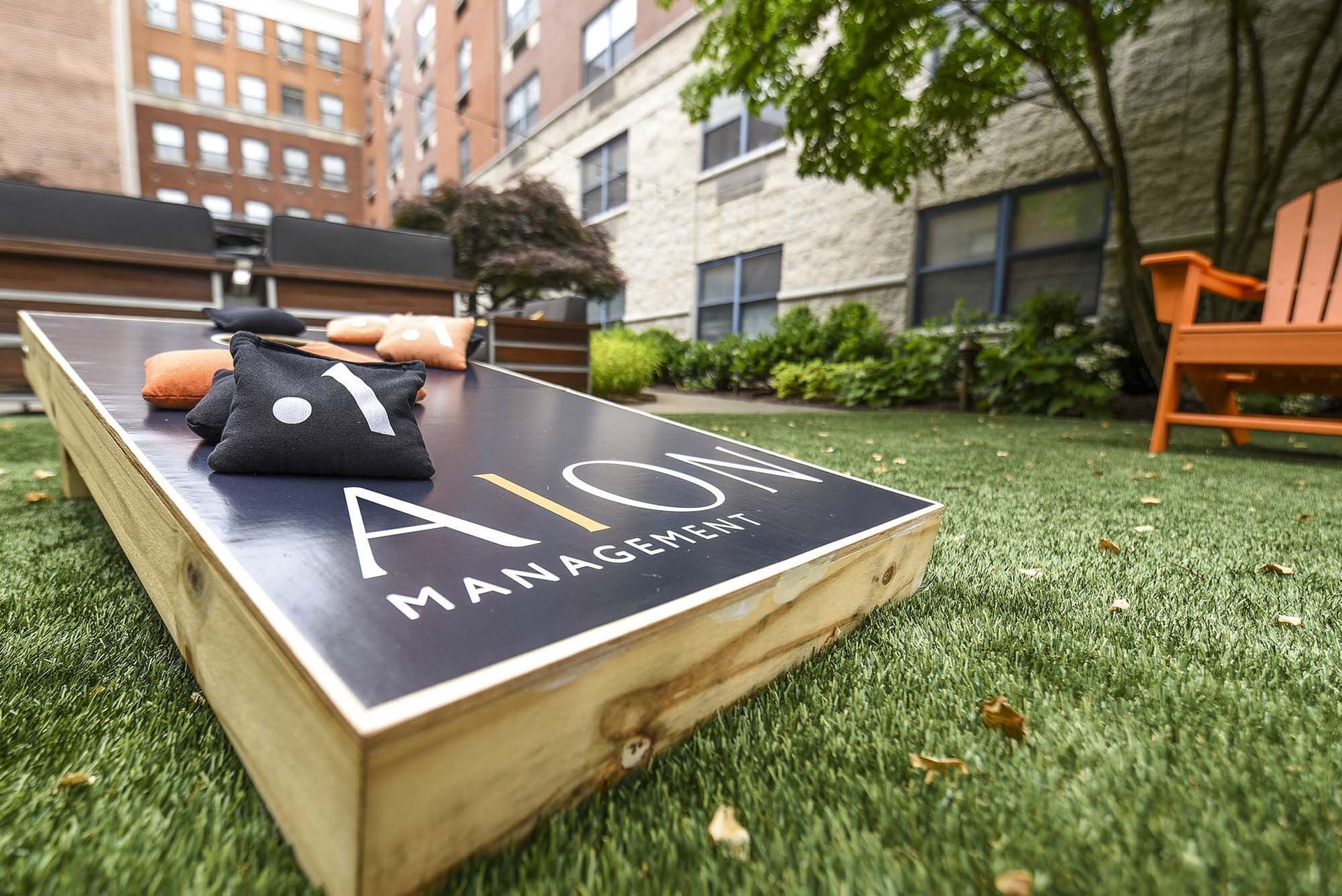 Cornhole game on lawn with bean bags, orange chair, and brick building background.